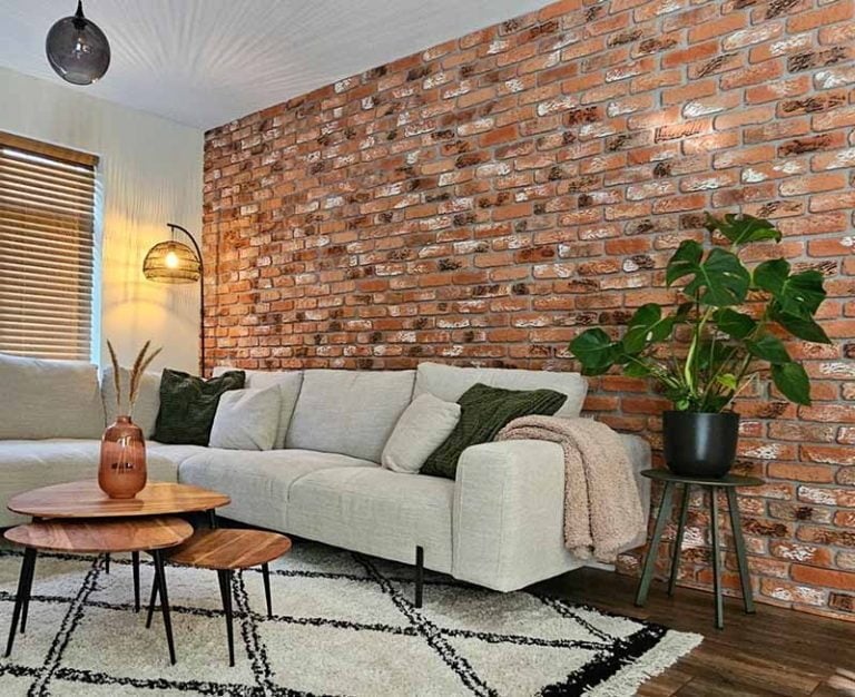 Woonkamer met baksteenstrips wandpanelen in roodbruin met wit patina, achter lichtgrijze hoekbank met groene kussens en houten salontafels