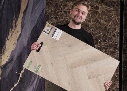 Een man glimlacht terwijl hij een groot houten vloer staal vasthoudt in een showroom, met op de achtergrond donkere marmerachtige texturen