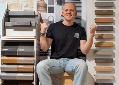 Man zittend tussen verschillende trapbekleding samples, glimlachend en met duimen omhoog, met stalen van hout en andere materialen in een showroomomgeving