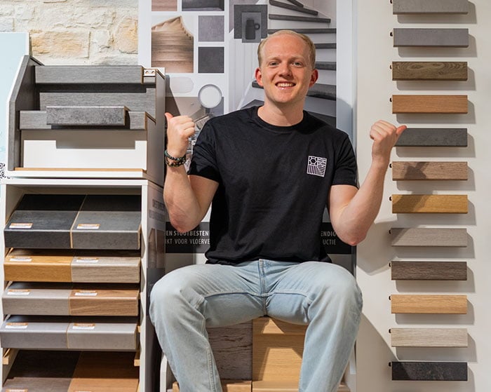 Man zittend tussen verschillende trapbekleding samples, glimlachend en met duimen omhoog, met stalen van hout en andere materialen in een showroomomgeving