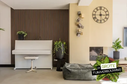 Moderne woonkamer met witte piano tegen een houten lattenwand, groene planten en wandklok, interieur door De Grote Tuinverbouwing