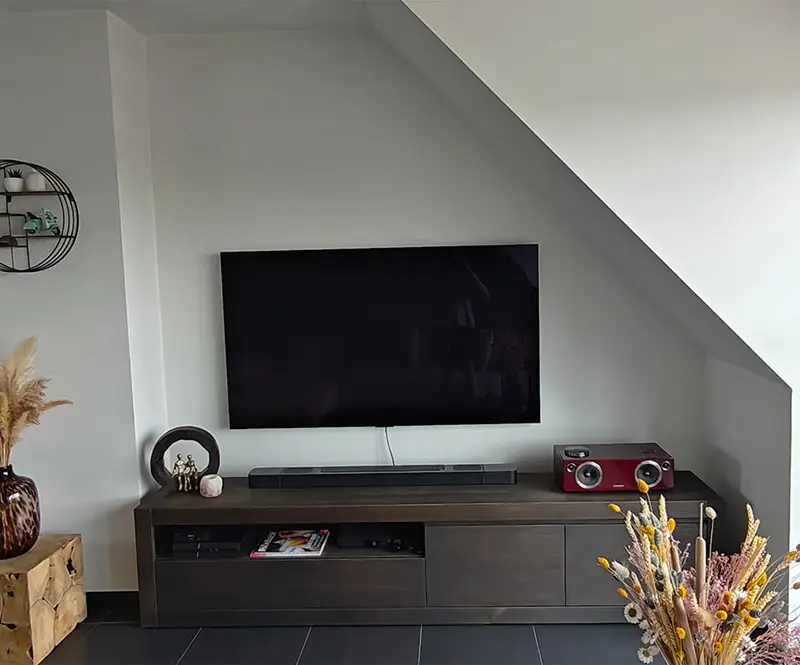 Woonkamerhoek met een tv aan een witte schuine wand boven een donker tv-meubel met decoratieve items