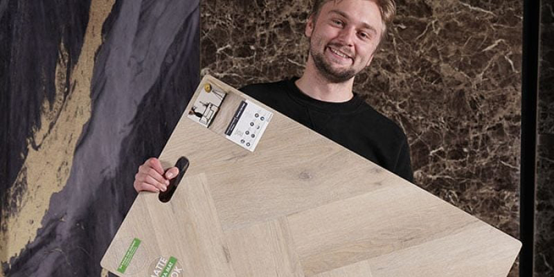 Een man glimlacht terwijl hij een groot houten vloer staal vasthoudt in een showroom, met op de achtergrond donkere marmerachtige texturen