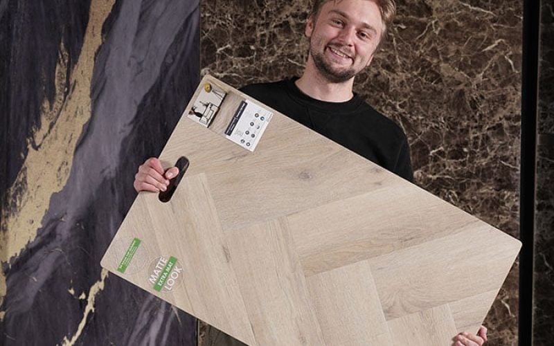 pvc vloer inspiratie Een man glimlacht terwijl hij een groot houten vloer staal vasthoudt in een showroom, met op de achtergrond donkere marmerachtige texturen
