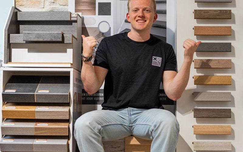 trap inspiratie Man zittend tussen verschillende trapbekleding samples, glimlachend en met duimen omhoog, met stalen van hout en andere materialen in een showroomomgeving