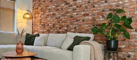 Woonkamer met baksteenstrips wandpanelen in roodbruin met wit patina, achter lichtgrijze hoekbank met groene kussens en houten salontafels