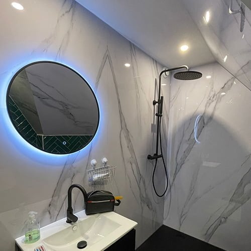 Badkamer met marmer wandpanelen ronde spiegel met verlichting zwarte wastafelkraan en douchekop